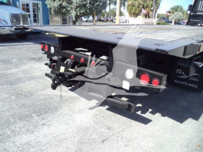 Flatbed trailer rear with dual red taillights and hydraulic/air connectors, Peterbilt 567 rollback truck seen in background, platform surface visible.