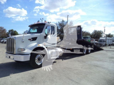 Peterbilt 567 tow truck with a flatbed rollback, white cab, black bed, in a lot.