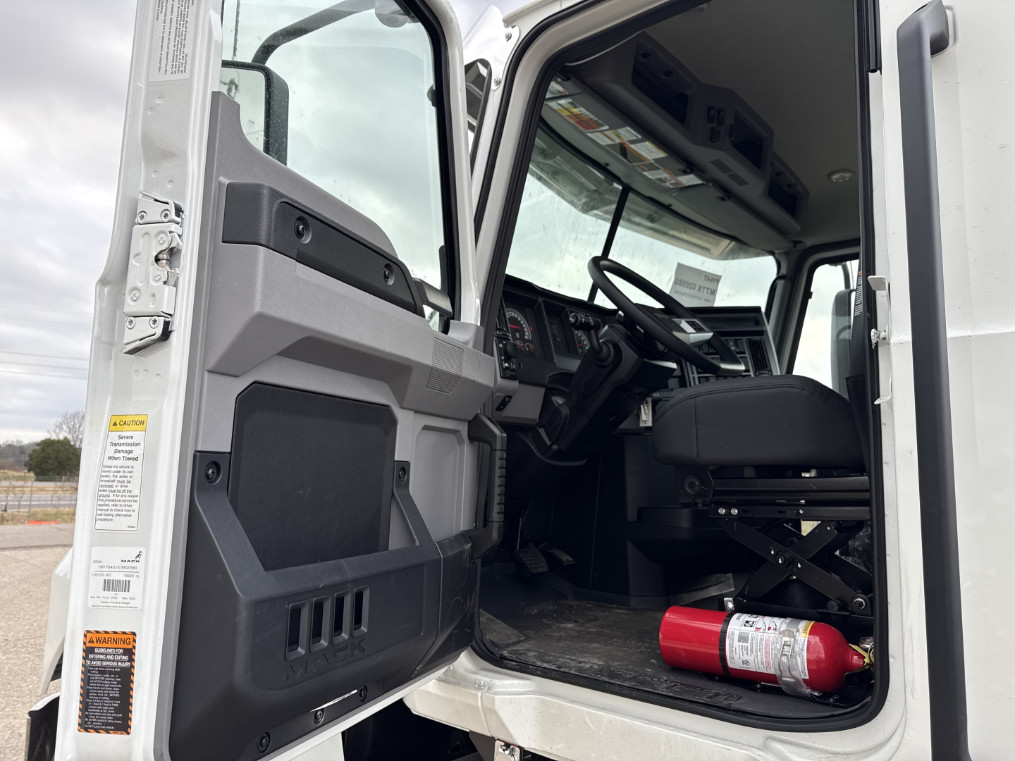 Truck cab interior of a new 2026 Mack PI64T with open door showing driver seat and dashboard, red fire extinguisher on floor, steering wheel visible.