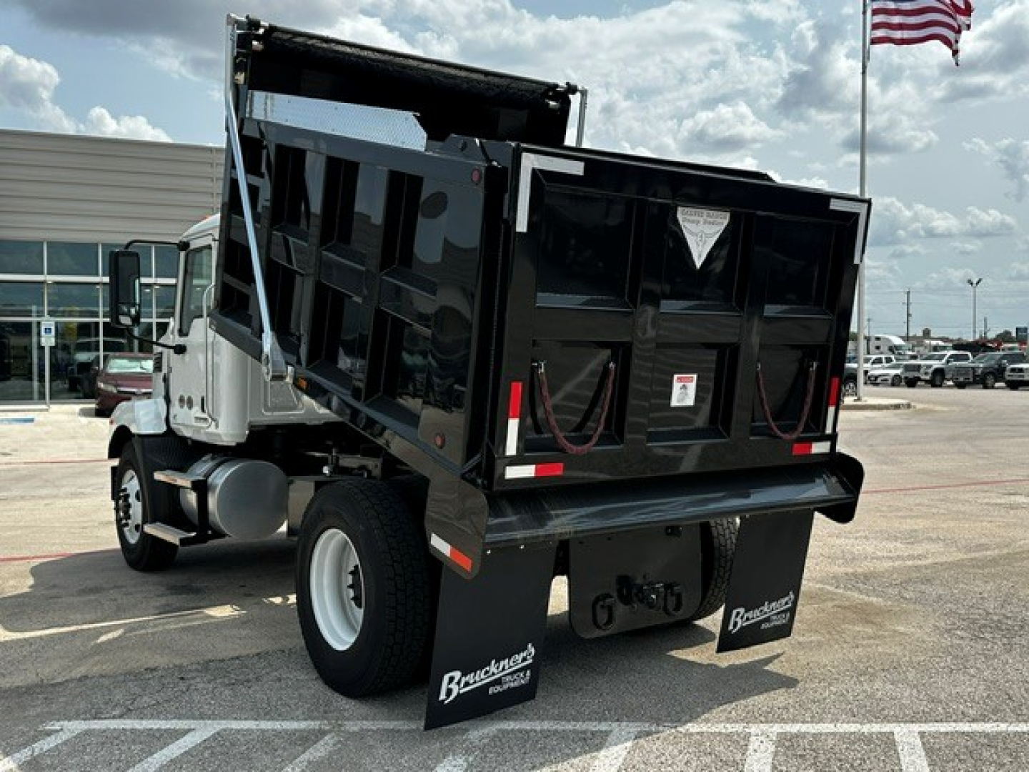 Dump truck rear view showing block dump bed on a Mack MD7 chassis with black hopper and mud flaps, white cab, dealership lot in background.