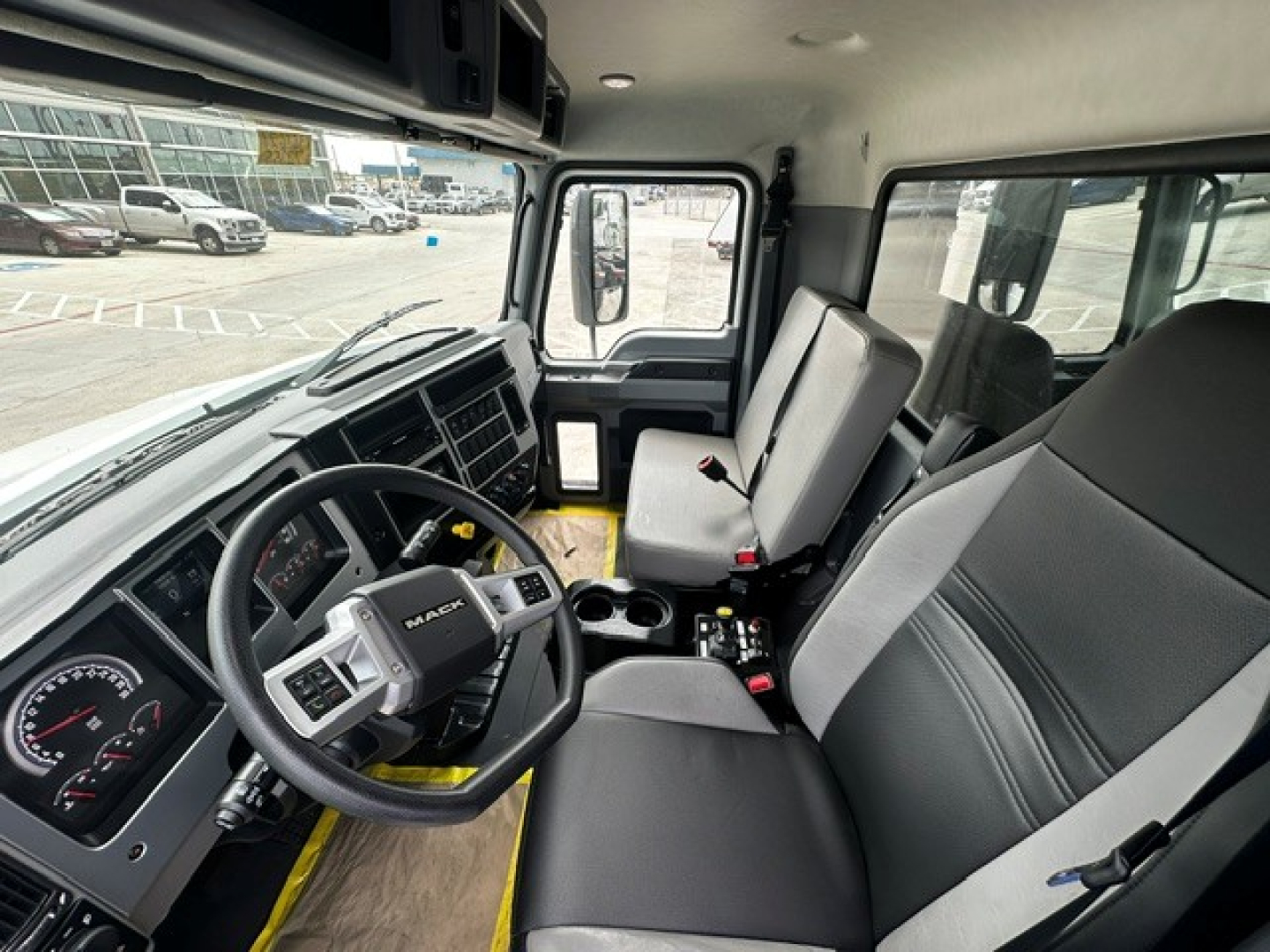 Cab interior of a 2027 Mack MD7 dump truck showing steering wheel, dashboard, driver seat, passenger seat, and center console with cup holders.