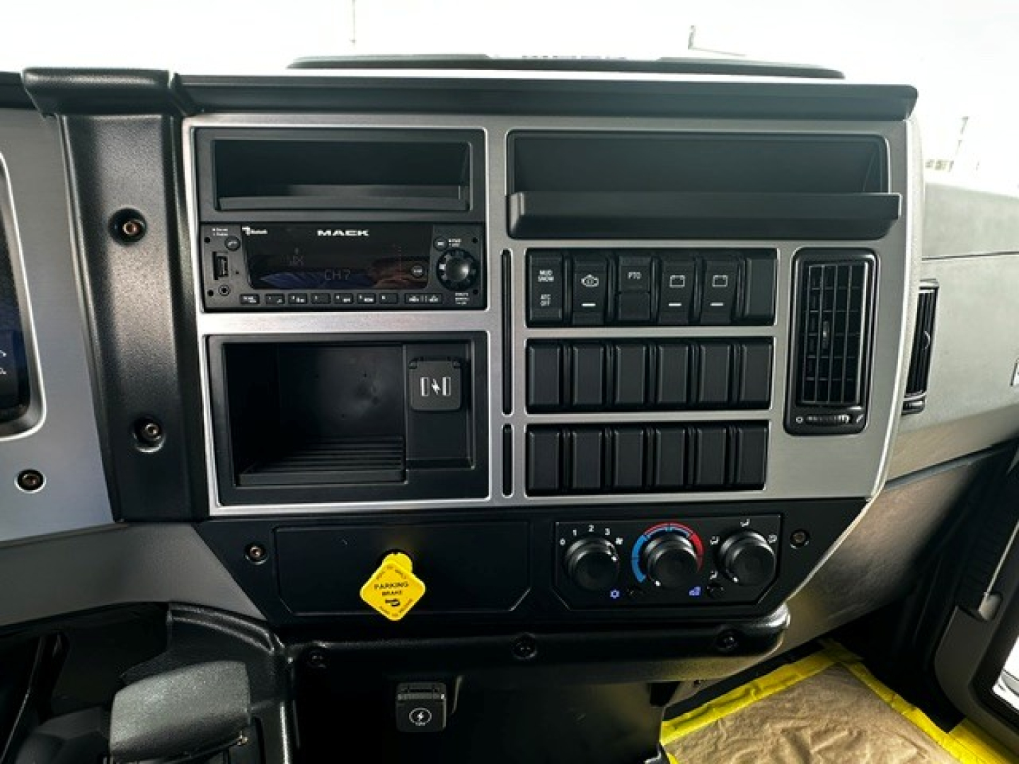 Dashboard of a Mack MD7 truck cab showing radio unit, climate controls, switch panel, and storage compartment with a yellow hazard tag.