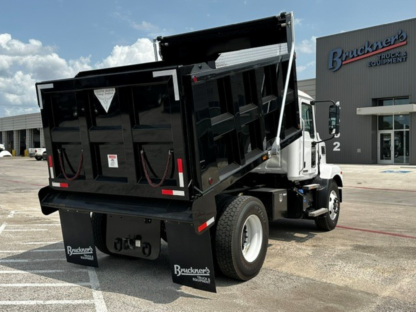 Dump truck rear/side view showing black dump body raised, Mack MD7 chassis, dual rear wheels, truck dealership in background.