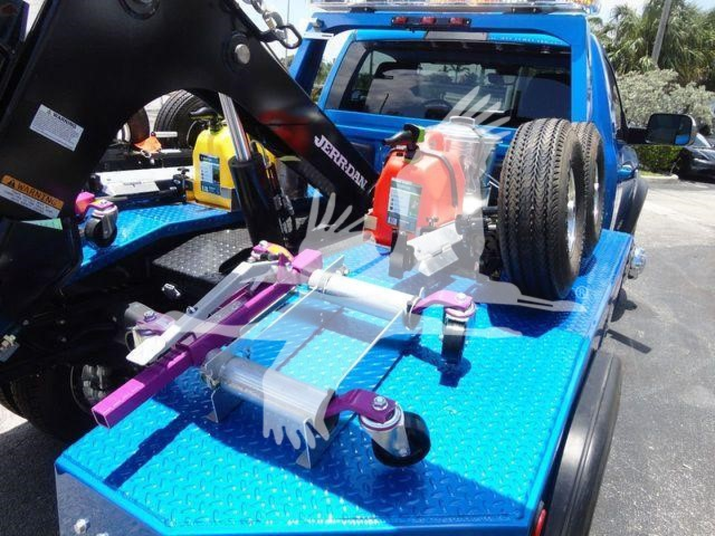Truck bed with winch equipment on blue flatbed, showing tow hooks, winch line hooks, and a spare tire mounted on the cab rear. J33417FM stock tag visible.