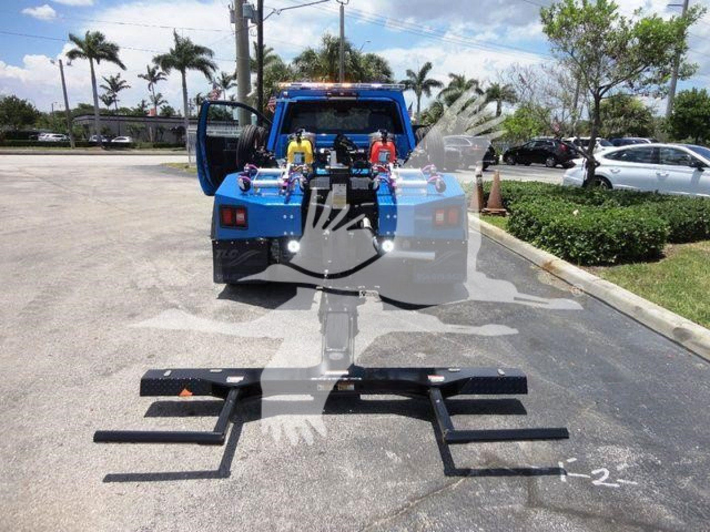 Ram 4500 tow truck rear with winch setup and tow bar components laid on pavement, blue body, open rear doors, palm trees in background.