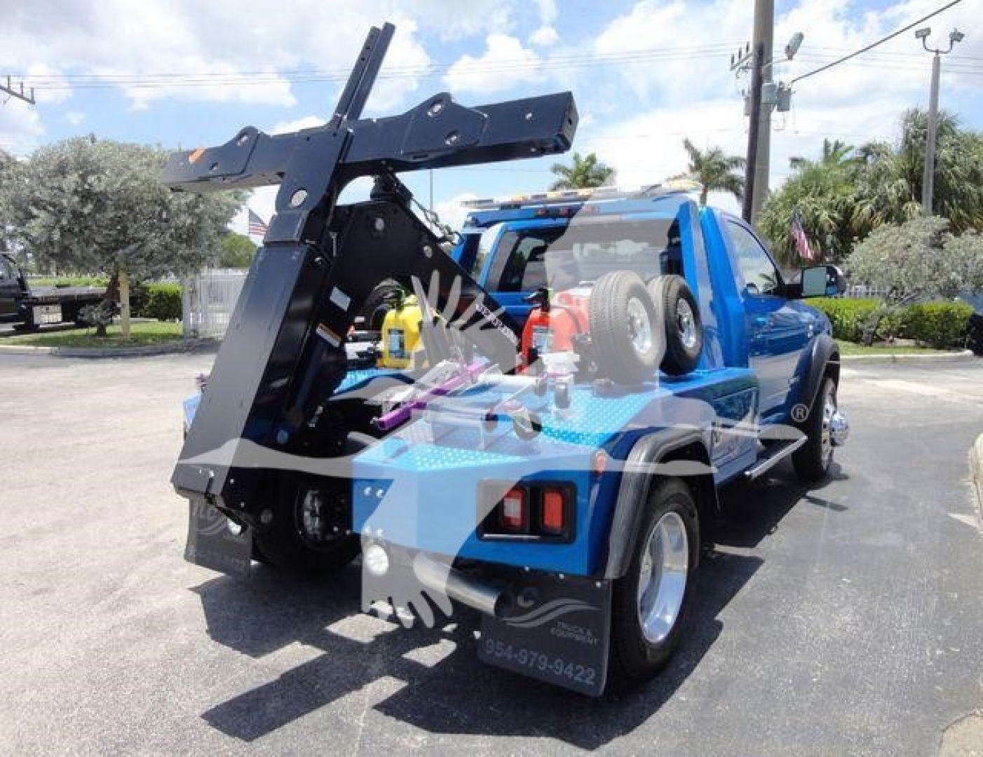 Blue Ram pickup truck with a mounted heavy-duty winch crane assembly in the truck bed and spare tire on the back, street scene in background.
