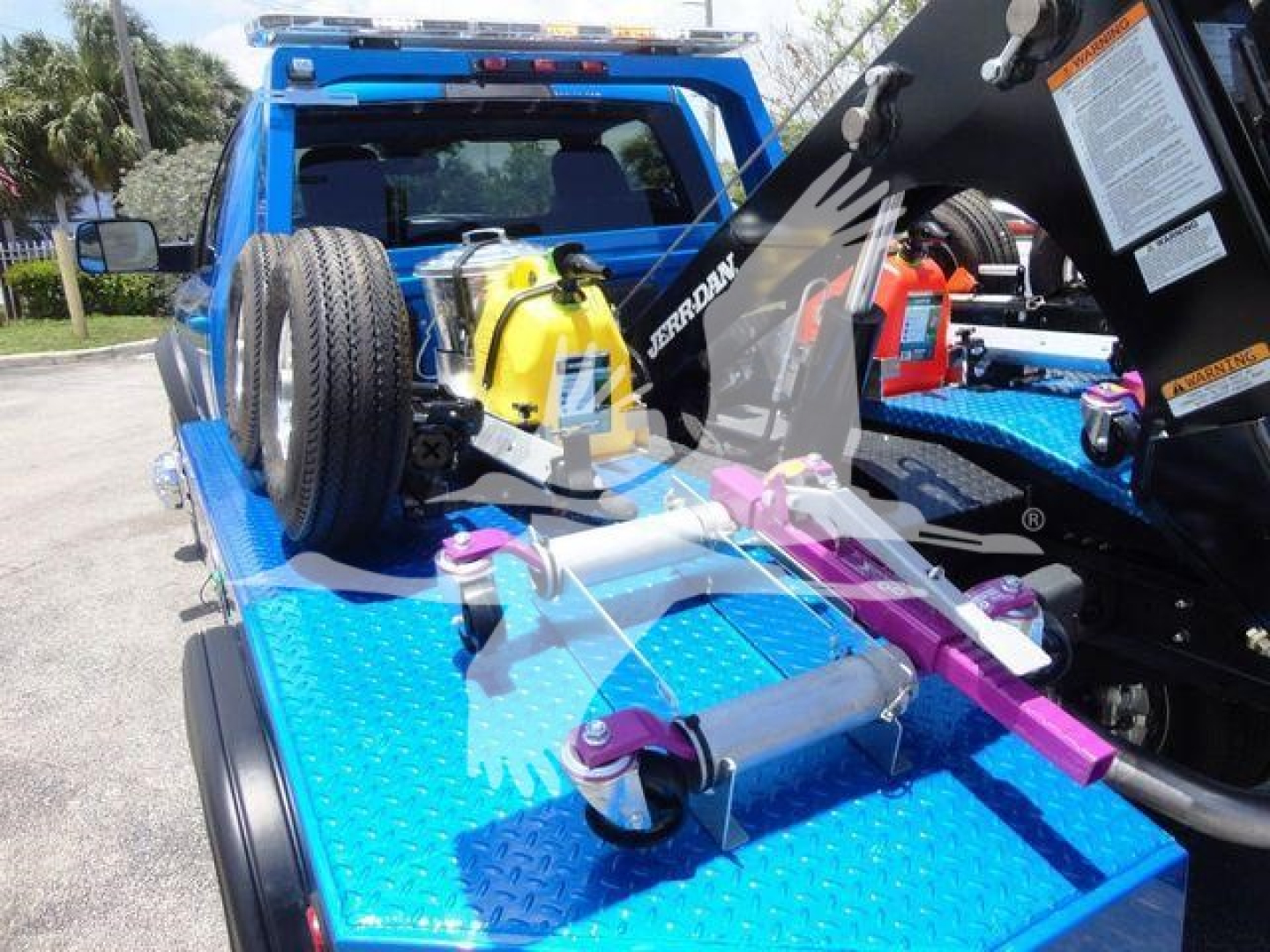 Ram 4500 tow truck bed with winch equipment and bright blue diamond plate bed; spare tire mounted, yellow portable fuel container, hydraulic winch lines, and control hardware visible.