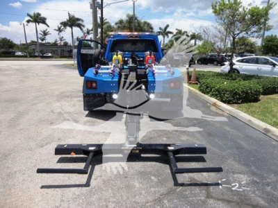 Ram 4500 tow truck rear with winch setup and tow bar components laid on pavement, blue body, open rear doors, palm trees in background.