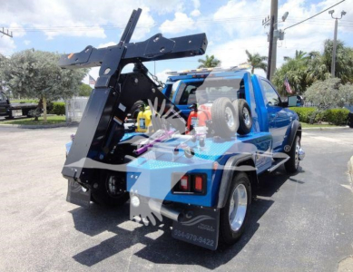 Blue Ram pickup truck with a mounted heavy-duty winch crane assembly in the truck bed and spare tire on the back, street scene in background.