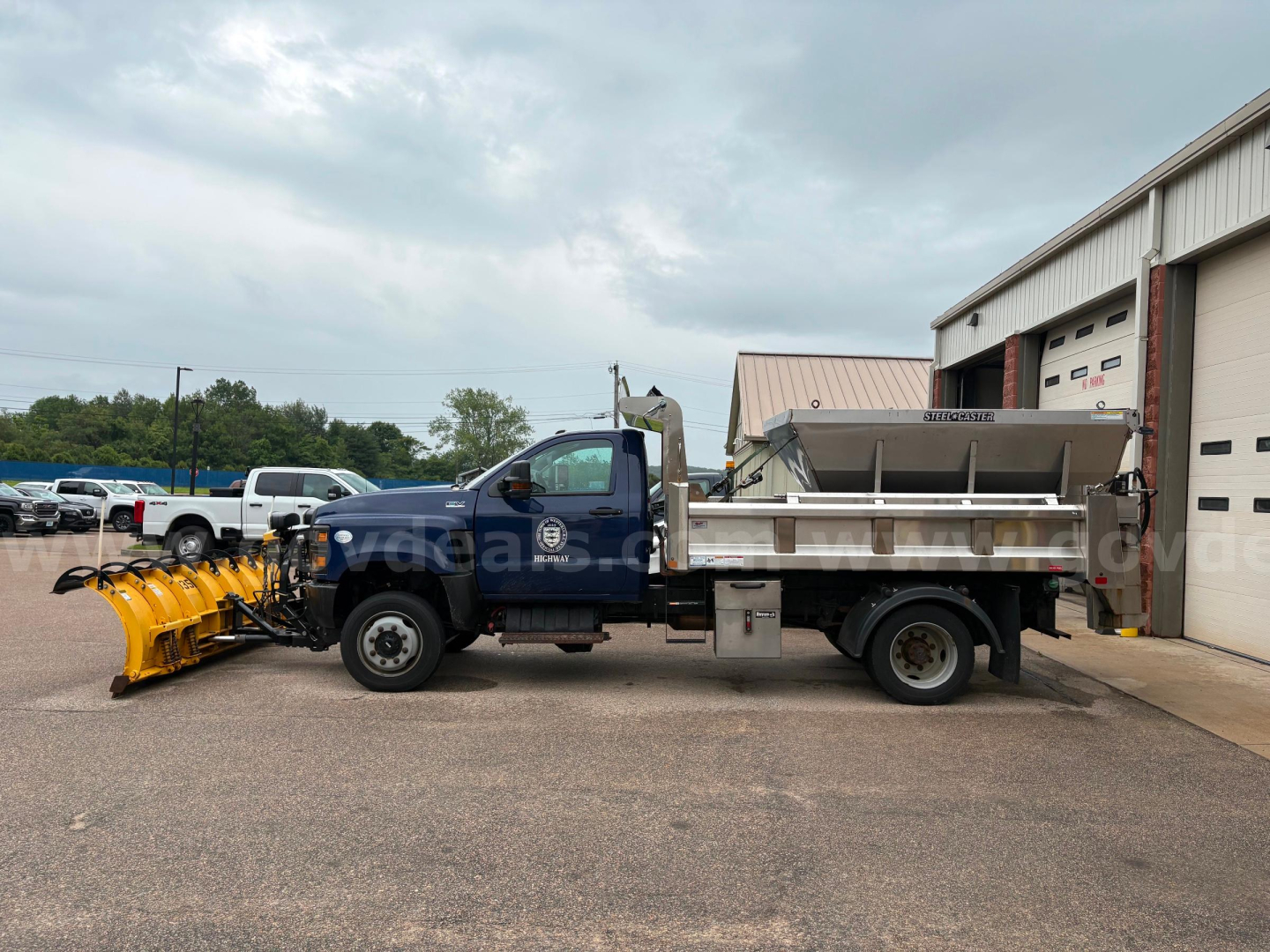 Dump truck side view showing front cab with blue paint and black grille, snow plow attachment at front, dump bed with reflective stripes, and workshop building in background.