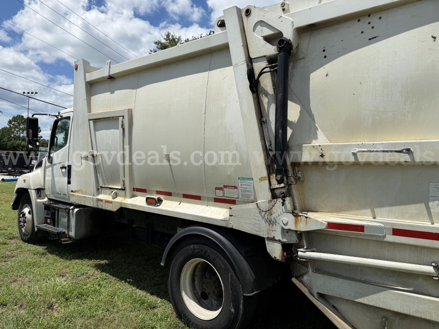 Truck side view of a 2015 Hino 338 refuse truck showing a large white dump body with hydraulic lift mechanism and safety decals on the side.