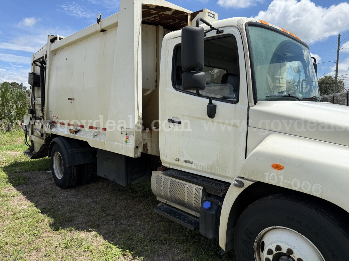 Garbage truck cab and rear body showing side mirror, door, wheel, and dump body with white paint and exposed hydraulic components.