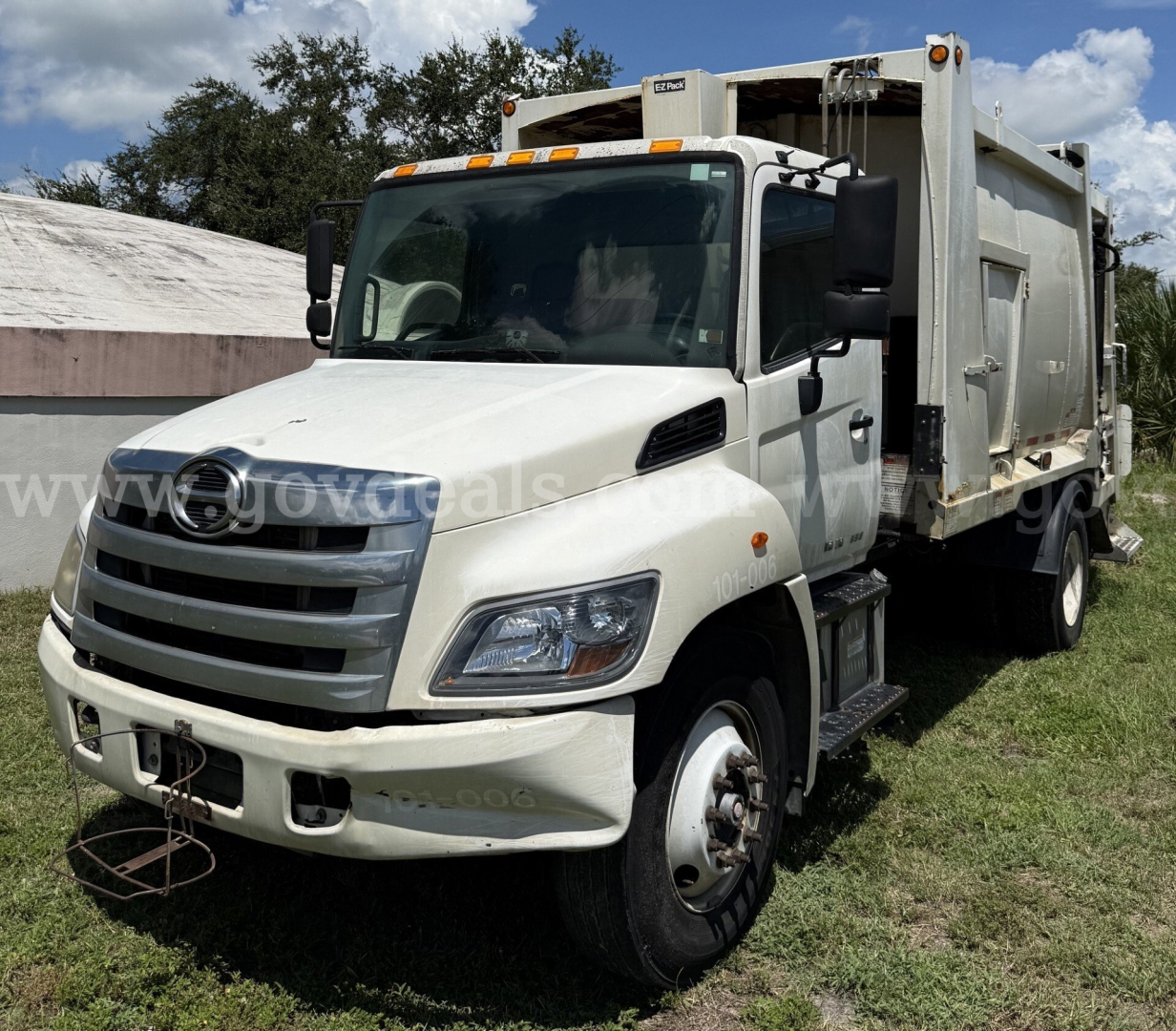White 2015 Hino 338 refuse truck in a grassy lot with a large rear dumpster body and side access steps.
