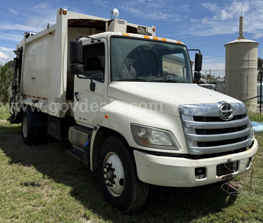 White 2015 Hino 338 garbage truck front-left view with cab and dump body, DOT lights on cab roof, grille, bumper, and dual rear wheels visible.