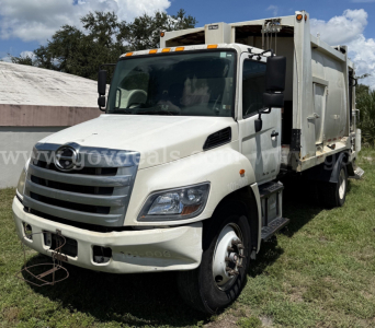 White 2015 Hino 338 refuse truck in a grassy lot with a large rear dumpster body and side access steps.