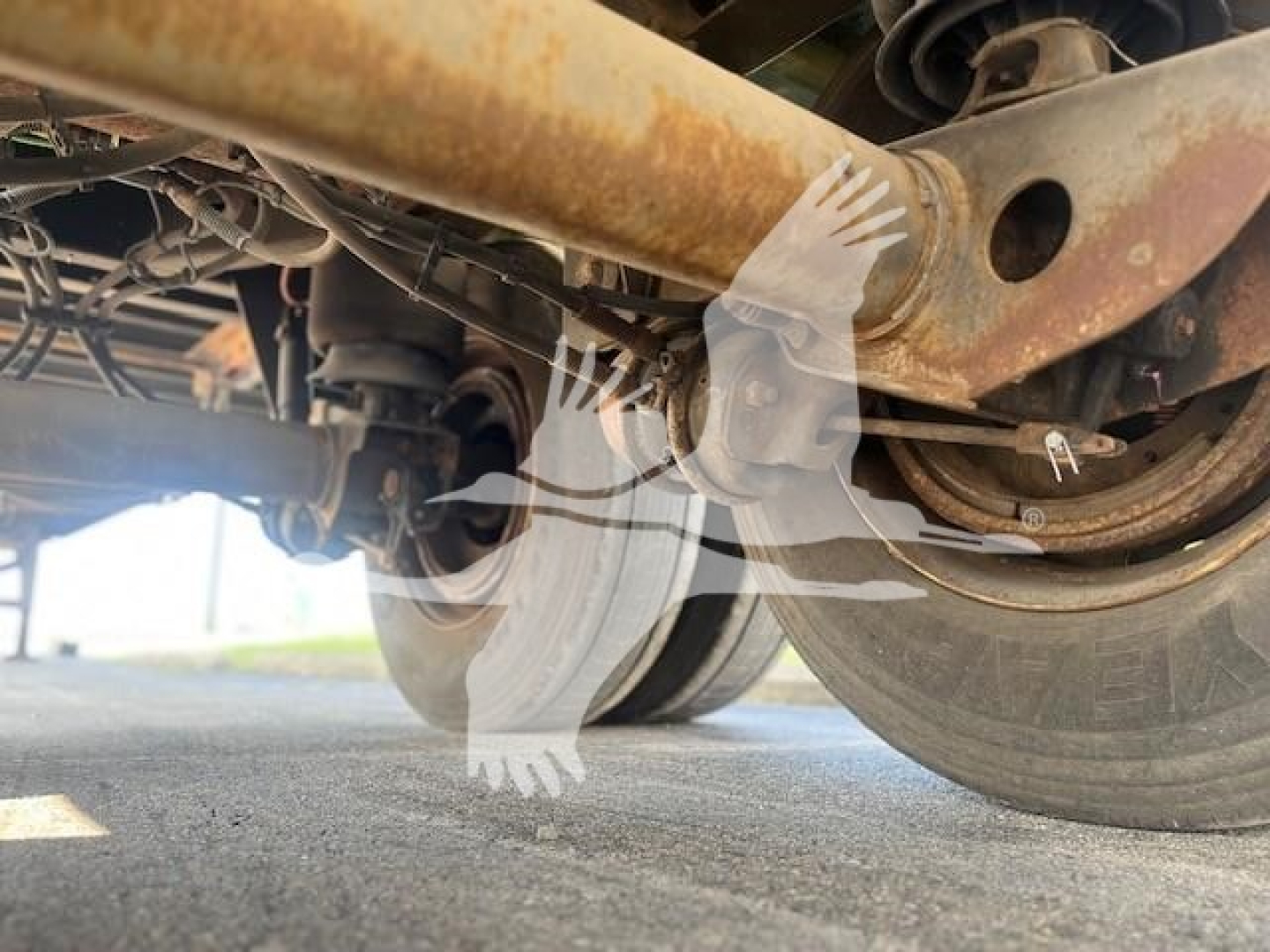 Trailer undercarriage with wheels and suspension, cylindrical axle beam, rust-colored metal components, and visible hub/axle assembly.