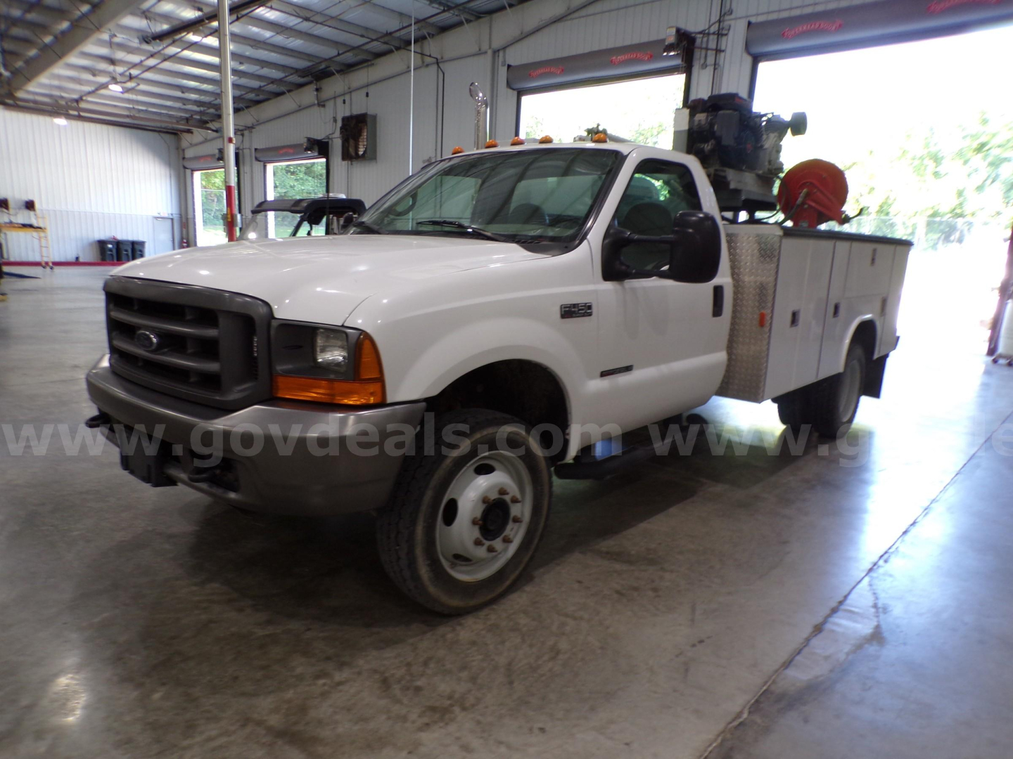 Ford F-450 utility truck in an indoor service bay showing front grille, single cab, service bed with equipment rack and hose reel.