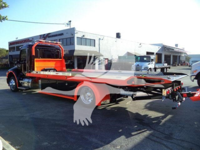 Kenworth T280 rollback tow truck chassis in a dealer lot, side view with flatbed, orange cab, hydraulic winch assembly visible.