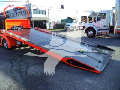 Kenworth T280 rollback tow truck platform with bright orange undercarriage and raised loading ramp in foreground; truck cabs in background.