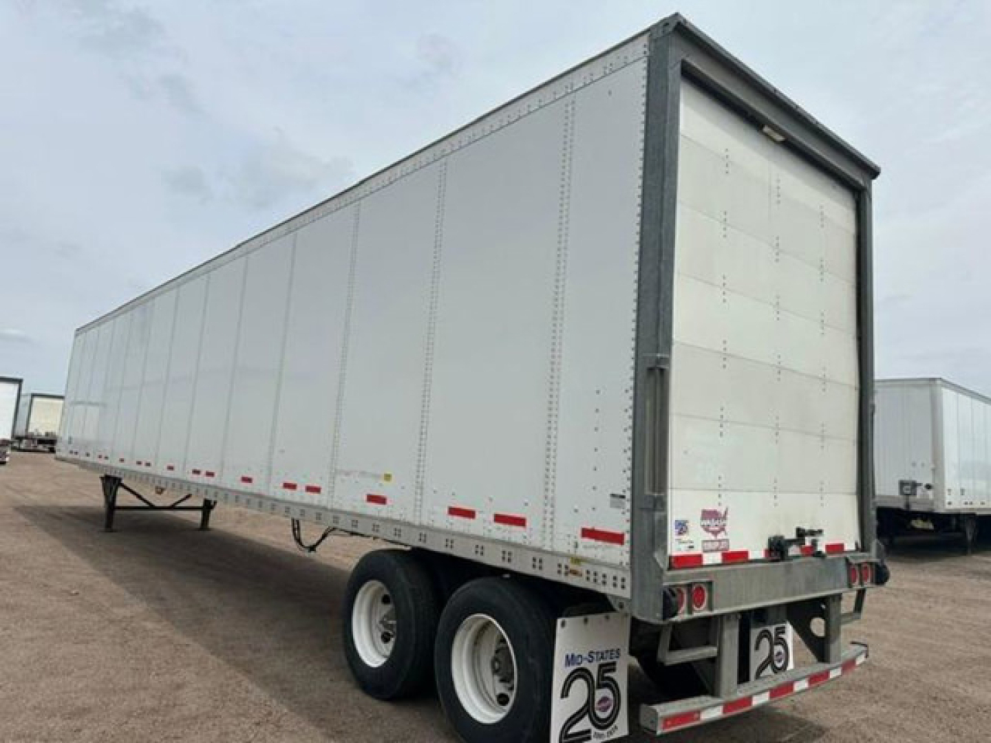 Trailer: white Wabash box trailer with double axles at rear, side height panels, rear doors. Visible markings include reflective tape along sides and rear, small yellow placards near the front, no visible text readable from this angle.