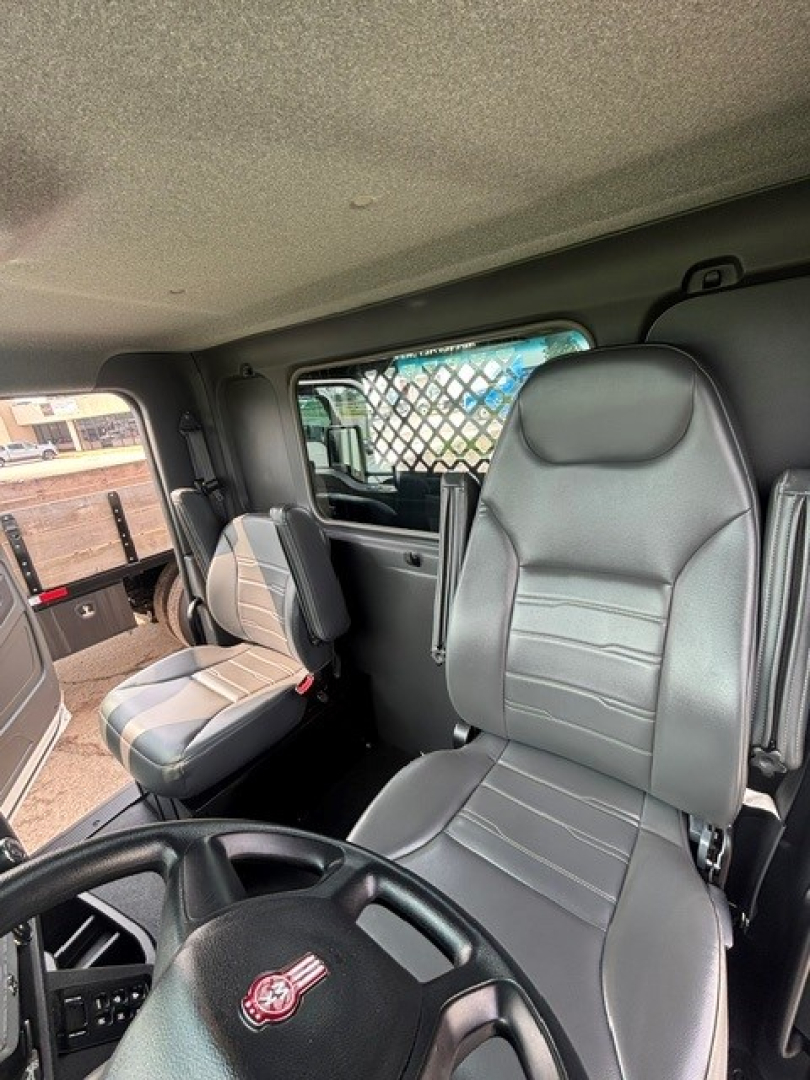 Cab interior of a Kenworth truck showing driver seat and passenger seat with gray upholstery, steering wheel with emblem, side window, and partition