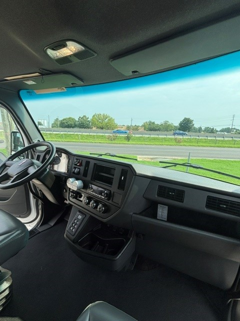 Cab interior of a Kenworth truck showing dashboard with steering wheel, central control panel, and gear selector. Windshield reveals highway view.