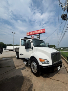 Kenworth T280 cab and chassis truck with open driver door, Kenworth sign in background, white cab, front grille and headlight details visible.
