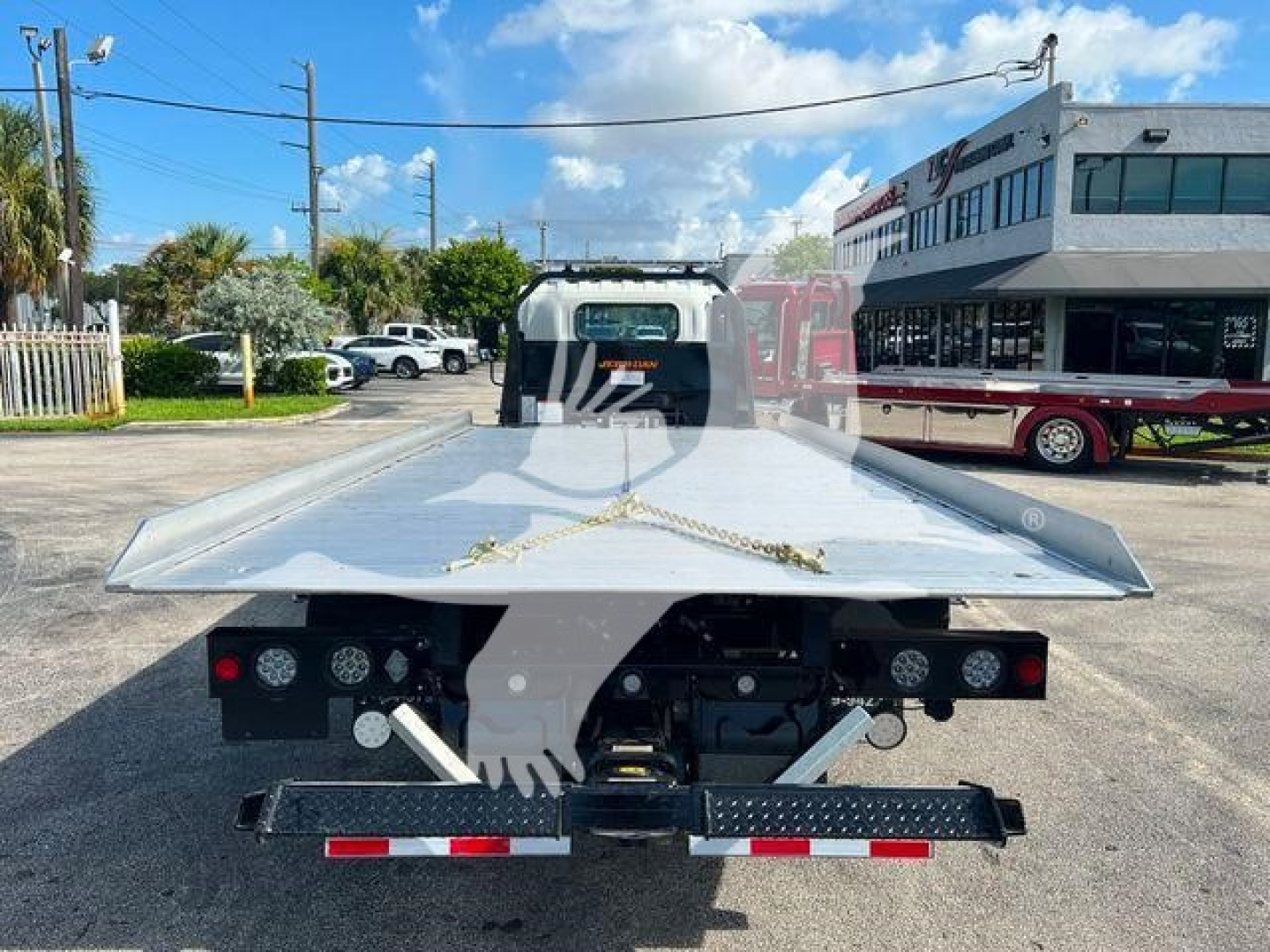 Isuzu NPR/NRR flatbed tow truck bed seen from rear, aluminum ramp bed with black bumper, multiple round taillights, and warning lights; chain tie-downs and equipment visible.