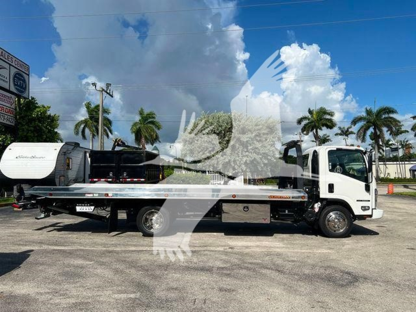 Isuzu NPR/NRR rollback tow truck side view showing flatbed with storage box and crane, white cab, blue sky with clouds.