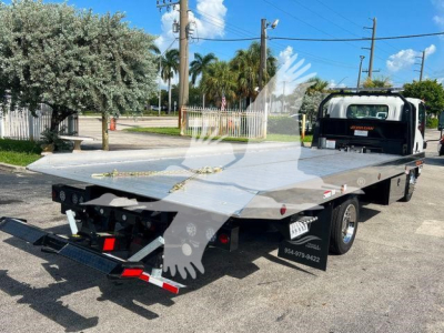 Isuzu NPRR rollback tow truck flatbed with metal deck and rear loading ramps, side rails, and dual rear wheels, viewed from rear-left in a parking lot.