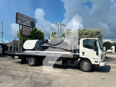Isuzu NPR/NRR series rollback tow truck in yard showing flatbed with crane, white cab, and dealer signage in background.