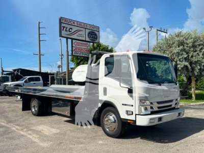 Isuzu NPR rollback tow truck in a dealership lot showing a flatbed with TLC Truck Sales Center sign in the background.