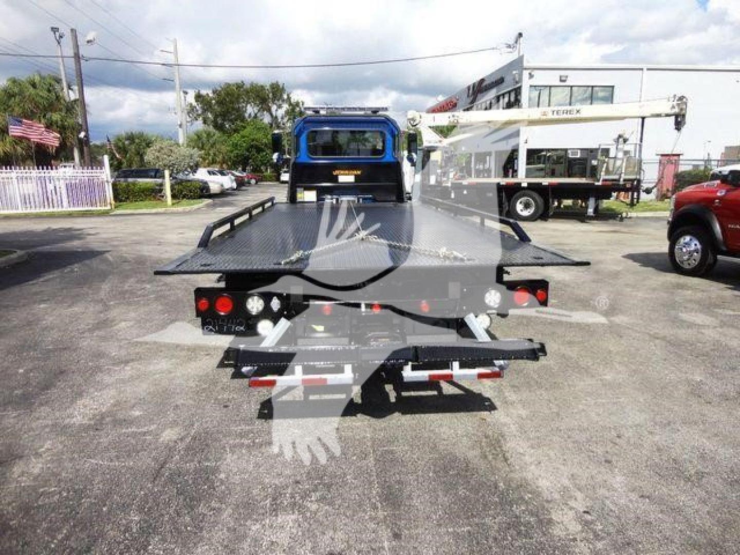 Truck rear with rollback flatbed, showing tail lights, tow hitch, and platform; crane in background; no legible model text visible.