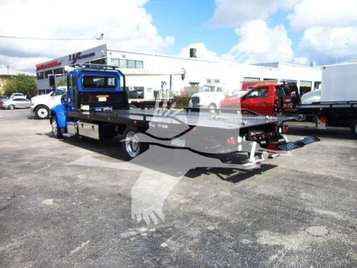 Peterbilt 337 rollback tow truck in blue with a hydraulic flatbed, side compartments, and winch in a dealership lot.