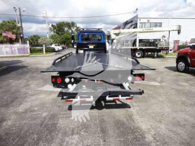 Truck rear with rollback flatbed, showing tail lights, tow hitch, and platform; crane in background; no legible model text visible.