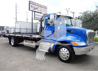 Blue Peterbilt 337 rollback truck seen from side, with flatbed, chrome front bumper, dual rear axles, and TLC Truck Sales Center sign in background.