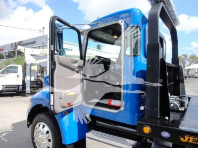 Truck cab side with open door, blue Peterbilt 337 rollback tow truck showing interior doorway, steps, and hydraulic equipment in rear.