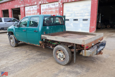 Chevrolet Silverado 2500HD crew cab flatbed truck with plow in a yard, green paint, flatbed rusty metal bed, dual rear wheels, utility equipment in background.
