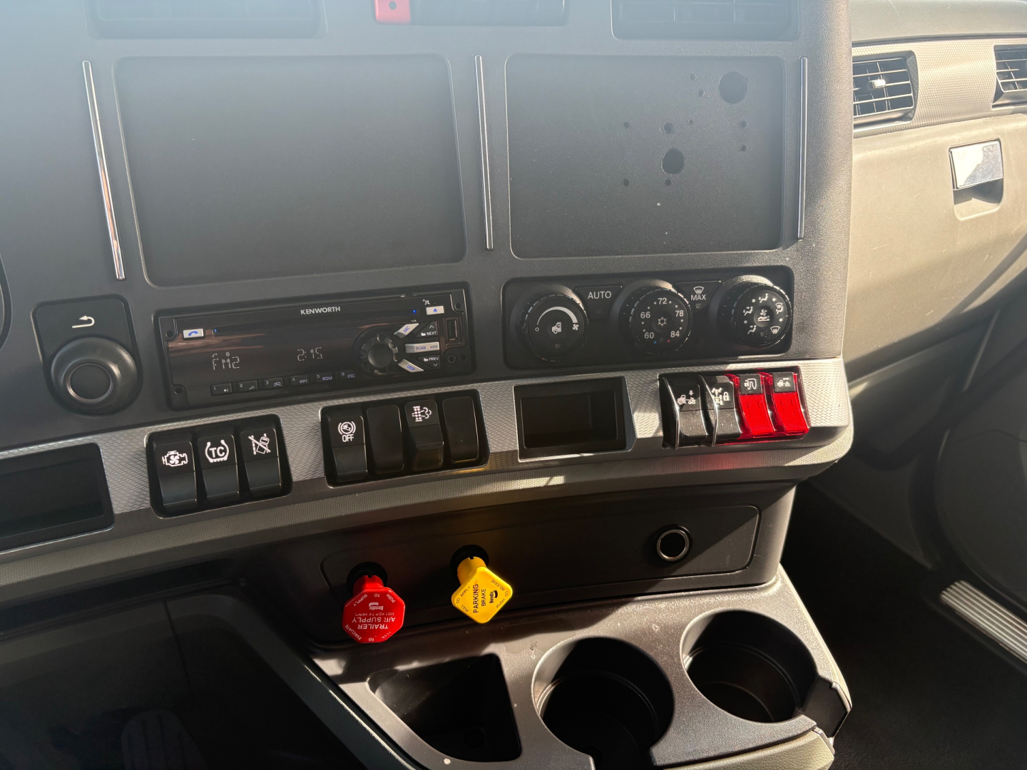 Kenworth T680 dashboard showing center console with radio, climate controls, and two colored knobs (red and yellow) with cup holders below.