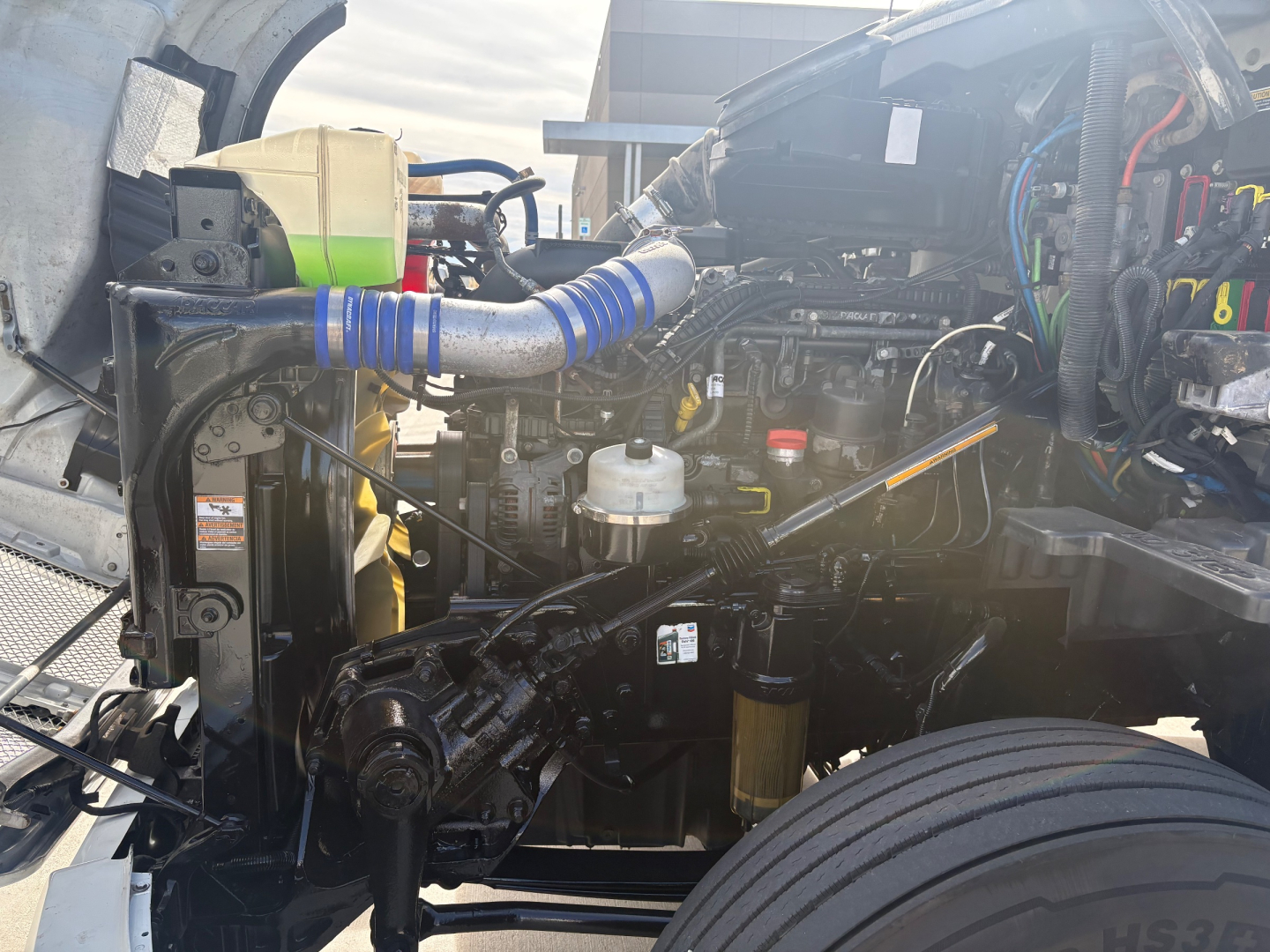 Kenworth T680 engine bay showing coolant reservoir, blue silicone hose, aluminum piping, and various hoses and wiring harnesses.