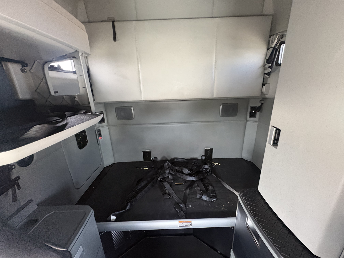 Interior sleeper cab of a truck showing a bed area with seat belts and a metal partition behind, grey panels, and storage compartments.
