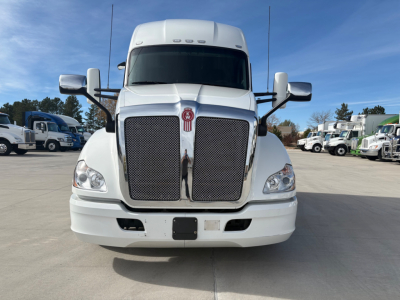Kenworth T680 front view showing large chrome grille, headlights, white cab, side mirrors, and air intake; image includes other trucks in background.