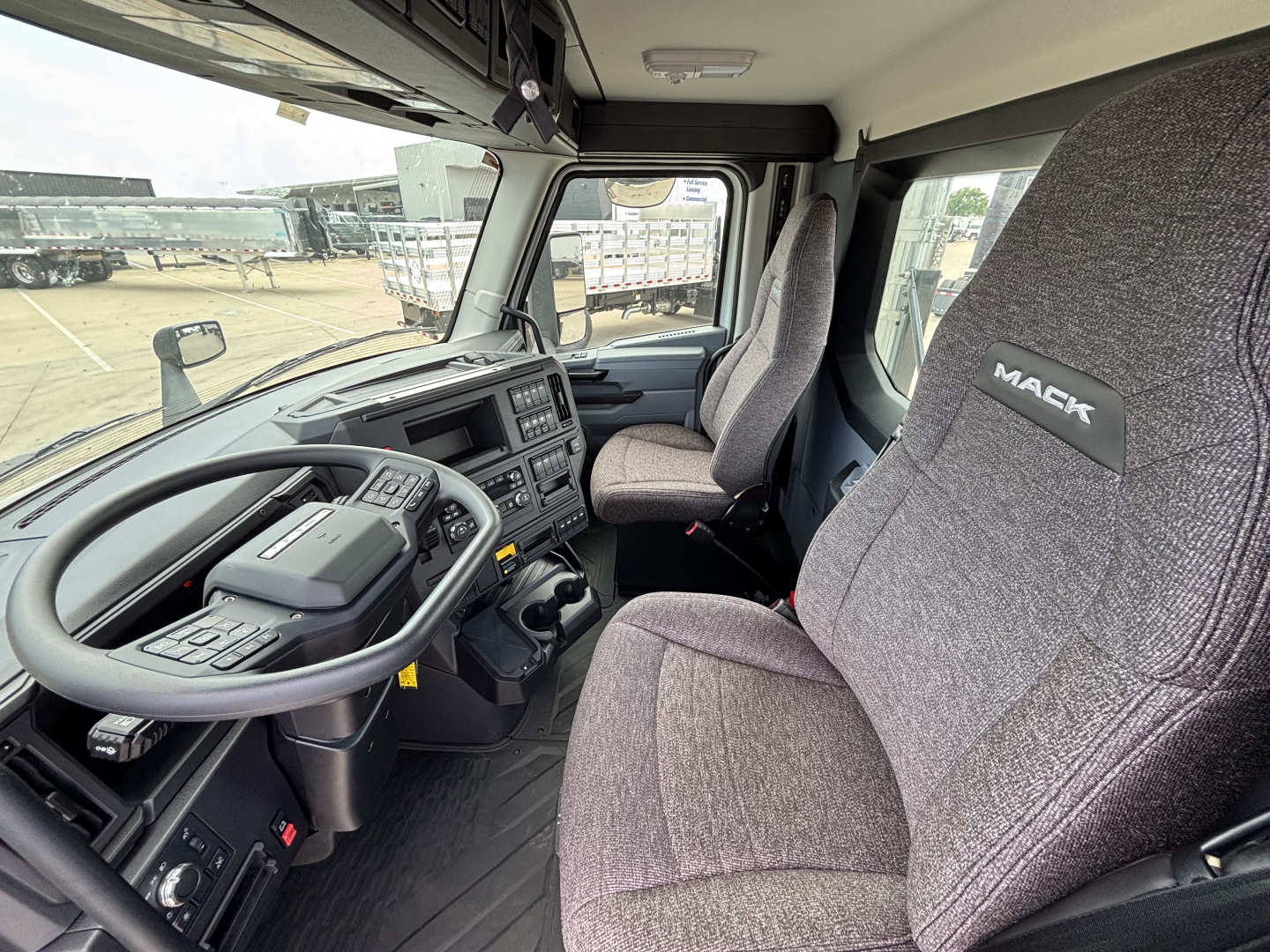 Truck cab interior showing steering wheel, dashboard, and gray fabric seats with MACK logo on the seat. Multiple controls on dash and center console visible.