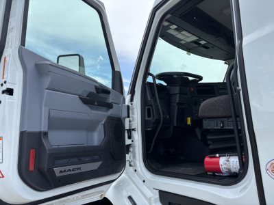 Truck cab interior of a 2027 Mack AN64 dump truck showing open driver door, steering wheel, dashboard, and a fire extinguisher on the floor near the door.