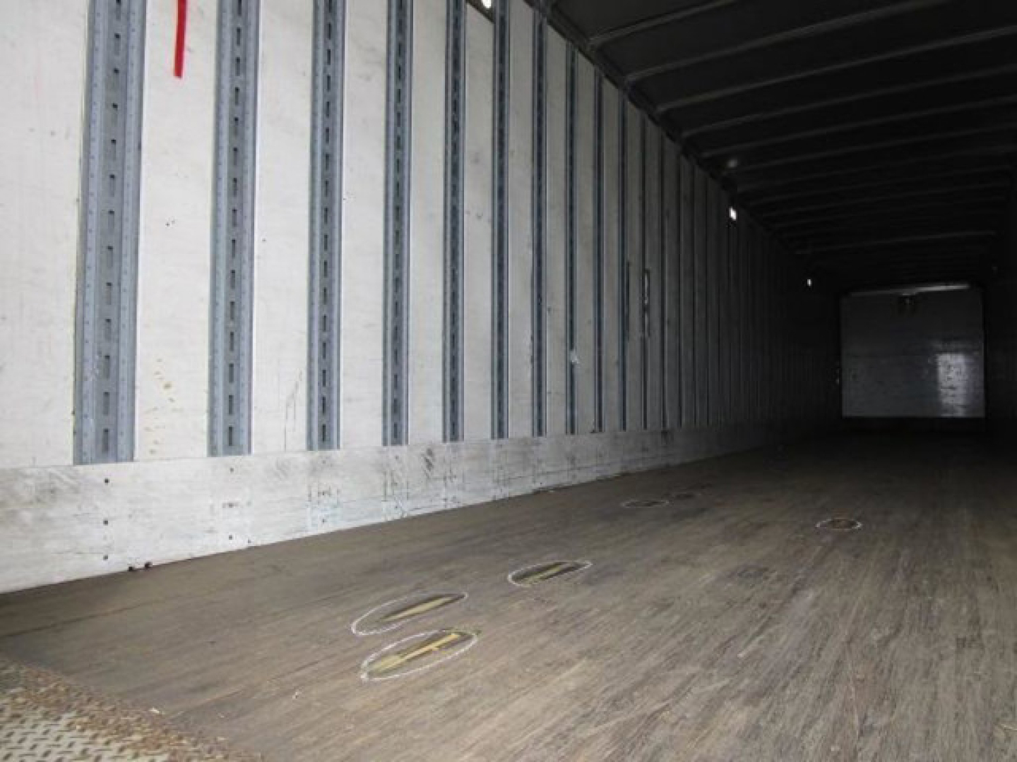 Interior of a cargo trailer with wooden floor and vertical steel ribs; circular tie-down rings on the floor; distant back wall appears dim.