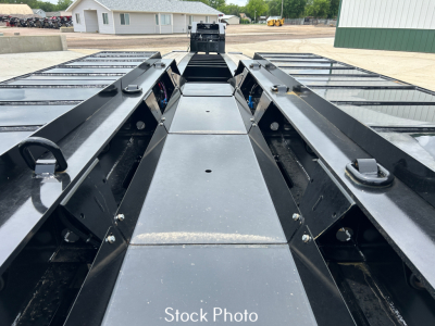 Truck trailer deck view showing long black metal frame rails, winches, and mid-section with sturdy side rails and gooseneck area; no general markings visible.