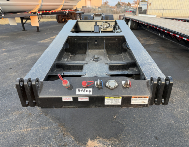 Load King trailer rear with hitch and multiple safety labels, including a red loop handle, and instructional stickers. The center shows a round carrier and metal frame with a long lowboy bed.