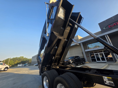 Dump truck chassis with raised dump bed showing frame and dual rear axles, Mack Granite likely; exterior building in background.