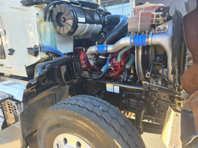  Mack Granite engine bay with visible coolant reservoir, air intake, hoses, and dual rear wheel in foreground.