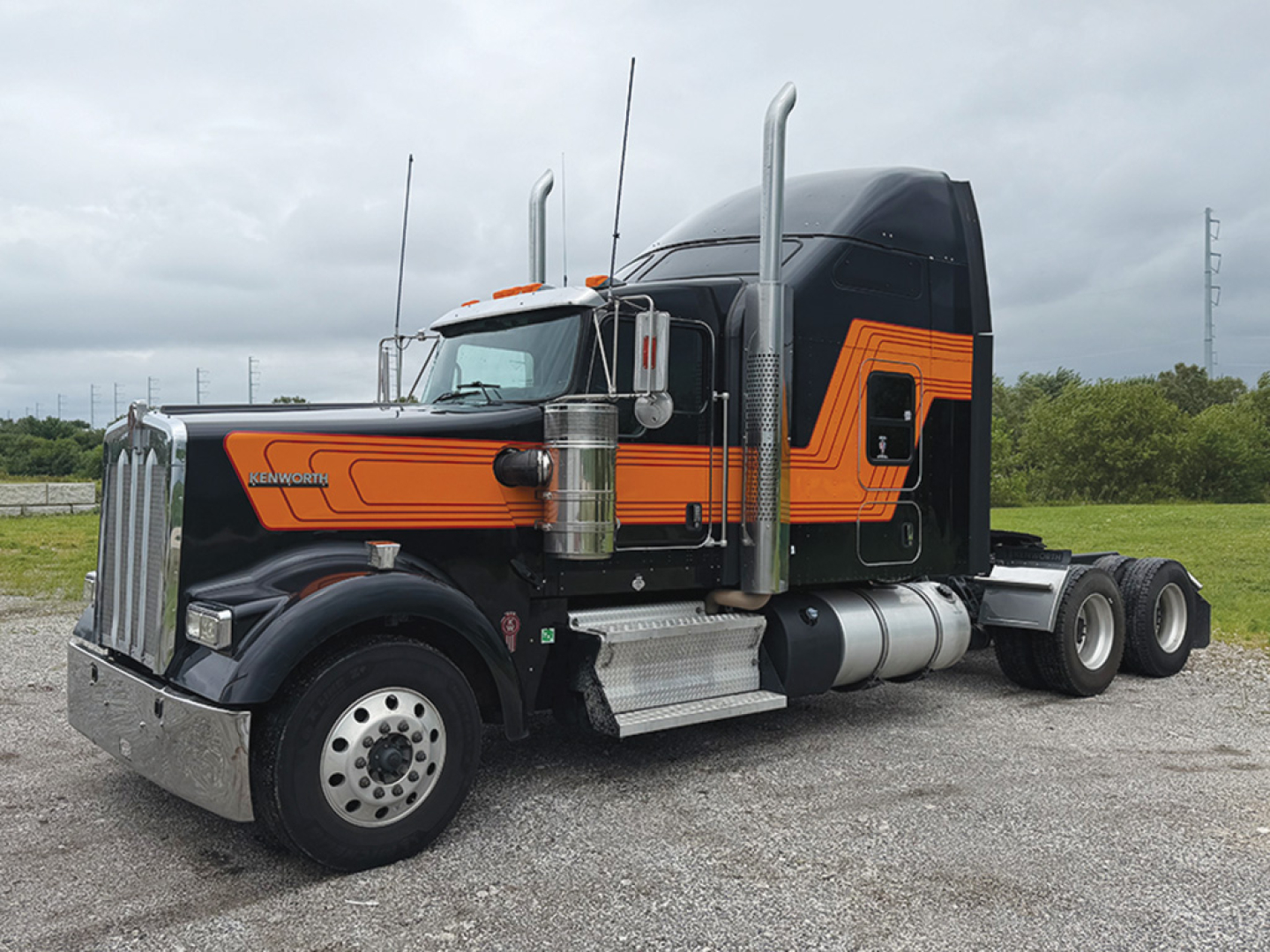 Kenworth W900L AeroCab tractor in profile showing orange and black paint, with chrome fuel tanks and dual exhaust stacks.